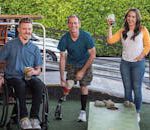 Three friends playing cornhole outdoors, enjoying drinks and smiling facing the camera. One is seated in a wheelchair, another is wearing shorts revealing a prosthetic leg.