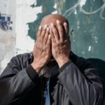 A bald man expresses frustration outdoors against a worn wall, covering his face with his hands