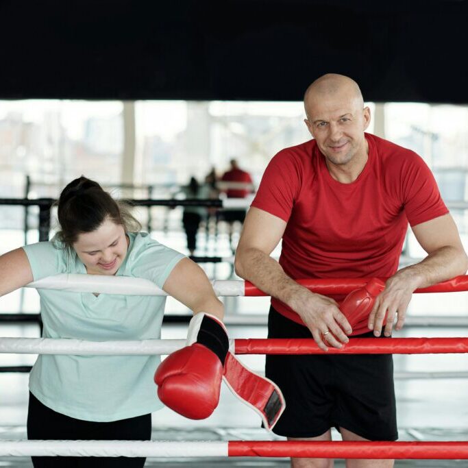 Male coach and woman with a disability wearing big boxing gloves about to climb in the rink for a friendly boxing session, emphasizing inclusivity and fitness.